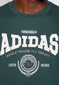 Primer plano de una camiseta Adidas de color verde oscuro con un logo blanco y negro, texto que celebra 50 años desde 1972 hasta 2022, y "Famosa en el Mundo por su Calidad."