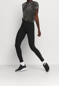 Persona vestida con leggings negros con logo lateral, calcetines blancos, zapatillas negras de Adidas y una camiseta de manga corta estampada frente a un fondo liso.