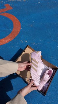 Tênis Adidas rosa em uma caixa de papelão, apresentando três listras douradas, um cabedal texturizado e uma sola de borracha. Forro de tecido macio visível.