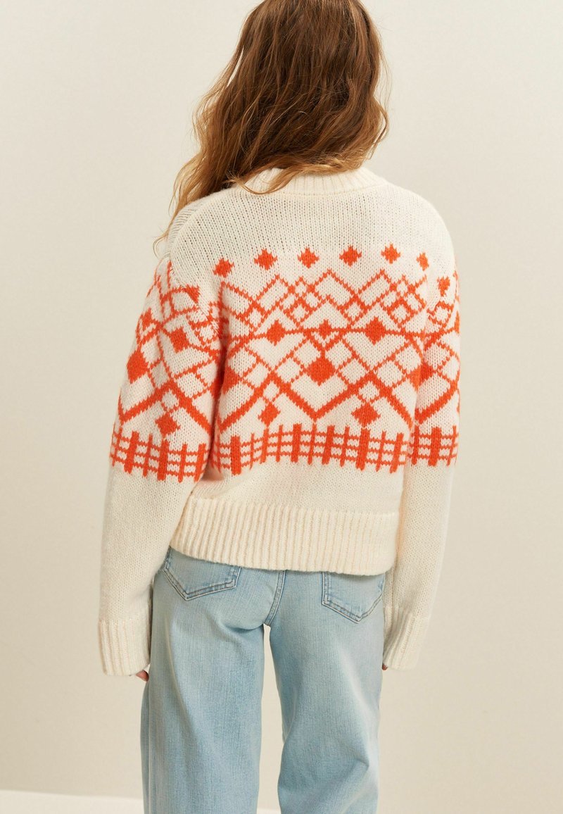 Cremfarbener Pullover mit einem orangefarbenen geometrischen Muster auf dem Oberkörper, ausgestattet mit gerippten Bündchen und Saum, über hellblauen Jeans getragen.