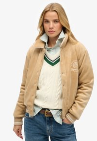 Beige corduroy jas met een rits en drukknoopsluiting, ribbelmanchetten en een geborduurd logo. Gekenmerkt over een roomkleurige kabelgebreide trui en een gestreepte blouse.
