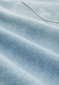 Helles blaues Denim-Gewebe mit weicher Textur und subtilen Farbvariationen, das eine Naht und ein Taschendetail aufweist.