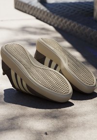 Paar sneakers met hak en witte zigzagzolen en bruine bovenkant, rustend met de zool naar boven op een zonovergoten betonnen oppervlak.