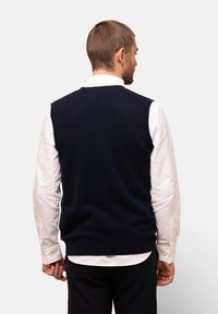 Marineblå stickad väst med rund halsringning, bärs över en vit knapp-up skjorta, som har en mjuk struktur och är ärmlös.