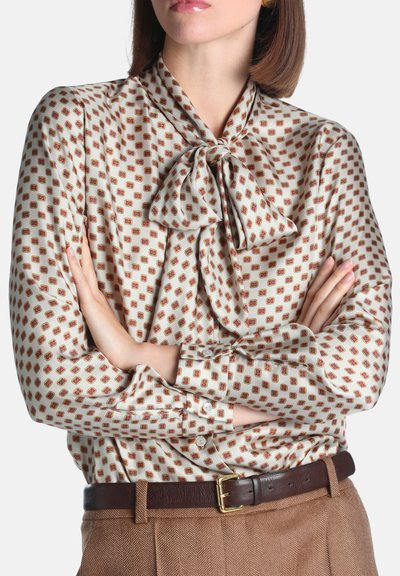 Femme, bras croisés, portant un chemisier à motifs avec un col lavallière, rentrée dans un pantalon marron taille haute et une ceinture en cuir foncé.