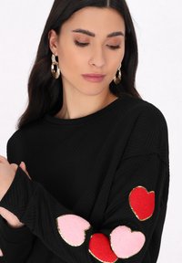 Zwarte lange mouw top met een getextureerd patroon, met roze, rode en rode vilt hart appliqués omrand met goud op de mouwen.