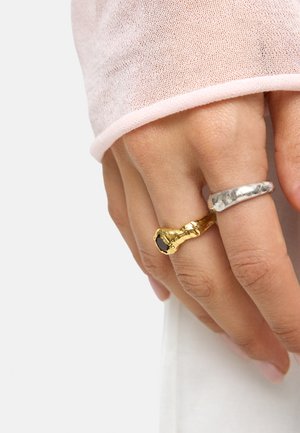 Ręka w jasnoróżowym rękawie nosząca dwa pierścionki: teksturowaną srebrną obrączkę oraz złoty pierścionek z czarnym kamieniem na palcach środkowym i serdecznym.