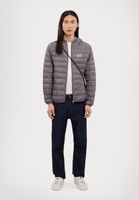 Grå pufferjakke med horisontalt quiltet mønster, lett glans, glidelås foran, båret over en hvit t-skjorte, kombinert med mørke jeans og hvite joggesko.