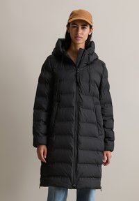Svart pufferjakke med hette og snøring, quiltet design, front glidelås og sidelommer, brukt over lyseblå jeans.