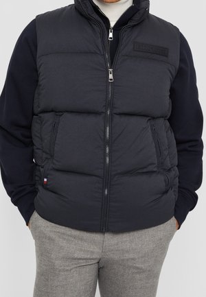 Mann trägt schwarze gesteppte Weste mit Reißverschluss über schwarzem Langarmshirt und grauer Hose, Hände in den Taschen, steht vor einfachem Hintergrund.