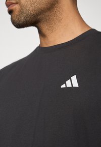 Közelkép egy férfiról, aki fekete pólót visel, fehér háromszög logóval a bal mellkas területén.