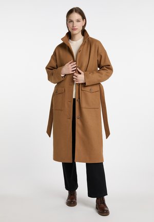 Jonge vrouw met een lange bruine jas met grote zakken, zwarte broek, beige trui en bruine veterschoenen, staand tegen een effen achtergrond.