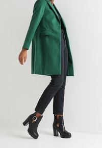 Abrigo verde de mezcla de lana con cierre de un solo botón y bolsillos laterales, combinado con vaqueros negros y botines negros con tacón y hebilla.