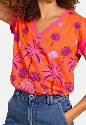 Woman wearing orange V-neck shirt with pink palm tree and seashell print and blue high-waist jeans, hand resting in pocket.