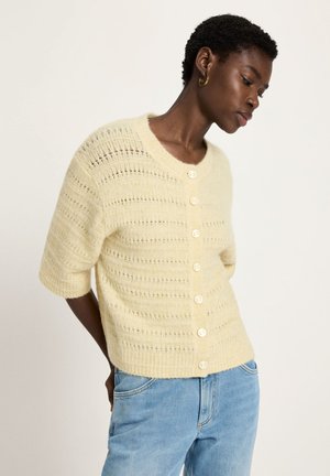 Person wearing a pale yellow short-sleeve knit cardigan with buttons and light blue jeans, posed with head tilted down and hands behind back.