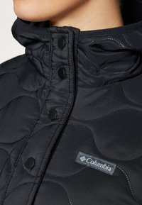Chaqueta acolchada negra con un cuello alto, botones de presión y una etiqueta del logo de Columbia. Exterior suave con un detalle de costura sutil.