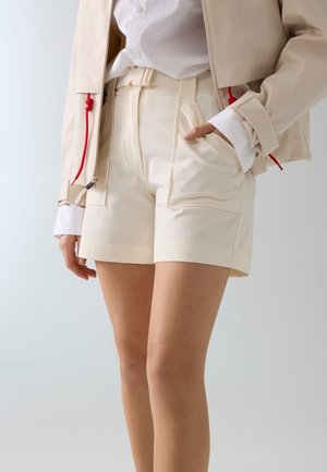 Vrouw draagt beige shorts met hoge taille en voorzakken, wit overhemd en beige jas met rode trekkoorden, staand met één hand in de zak.