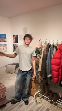 Νέος άνδρας στέκεται με καθημερινά ρούχα δίπλα σε κρεβάτι και βάση ρούχων με μπουφάν και μπότες σε ένα λιτό υπνοδωμάτιο.