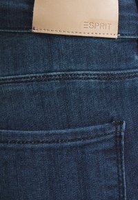 Donkerblauw denimstof met een zichtbare achterzak en een lichtbeige leren label met het merknaam "ESPRIT" geperst.