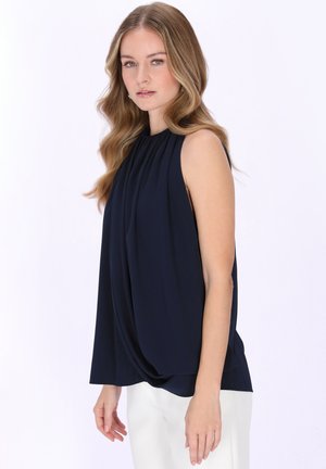 Młoda kobieta o falowanych blond włosach, ubrana w granatową, bezrękawnikową bluzkę z drapowaniem i białe spodnie, stojąca na tle jednolitego jasnego tła.