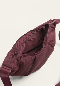 Bordo crossbody torba s glatkom teksturom, zatvorenim glavnim odjeljkom s patentnim zatvaračem i podesivim platnenim remenom. Lagana konstrukcija, obilje unutarnjeg prostora.