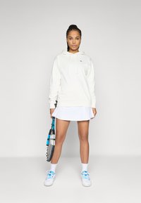 Hvit hettegenser med en frontlomme, snøring i hetta, og liten logo. Kombinert med et hvitt plisserte skjørt og blå detaljerte joggesko, mens hun holder en racket.