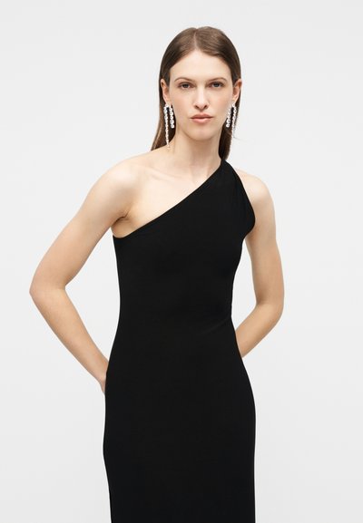 Γυναίκα που φοράει μαύρο φόρεμα με έναν ώμο, με μακριά ίσια μαλλιά και κρεμαστά σκουλαρίκια από κρύσταλλο, στέκεται μπροστά σε απλό λευκό φόντο.