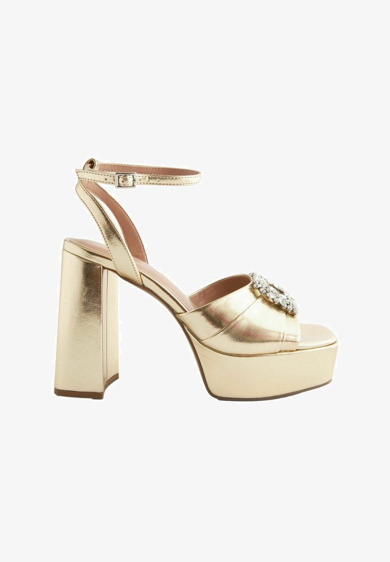 Sandali platform metallici dorati con un ampio tacco a blocco, caratterizzati da un design con cinturini incrociati e un accento di strass decorativo sulla parte anteriore.