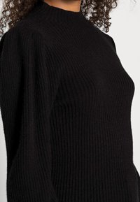Černý žebrovaný pletený svetr s vysokým límcem. Má dlouhé rukávy a přiléhavý střih, který zdůrazňuje texturu a tvar.