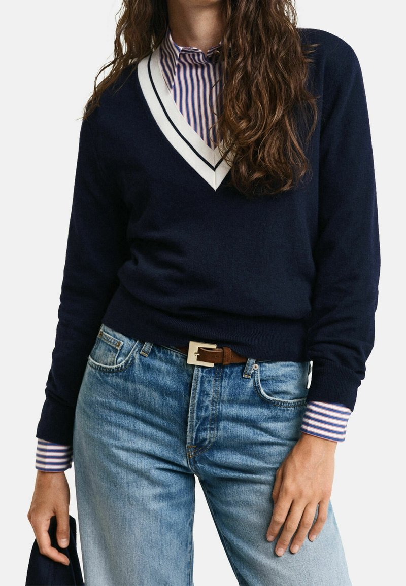 Personne portant un pull bleu marine à col en V avec bord blanc sur une chemise rayée, jean bleu clair avec ceinture marron, tenant un tissu sombre dans la main gauche.