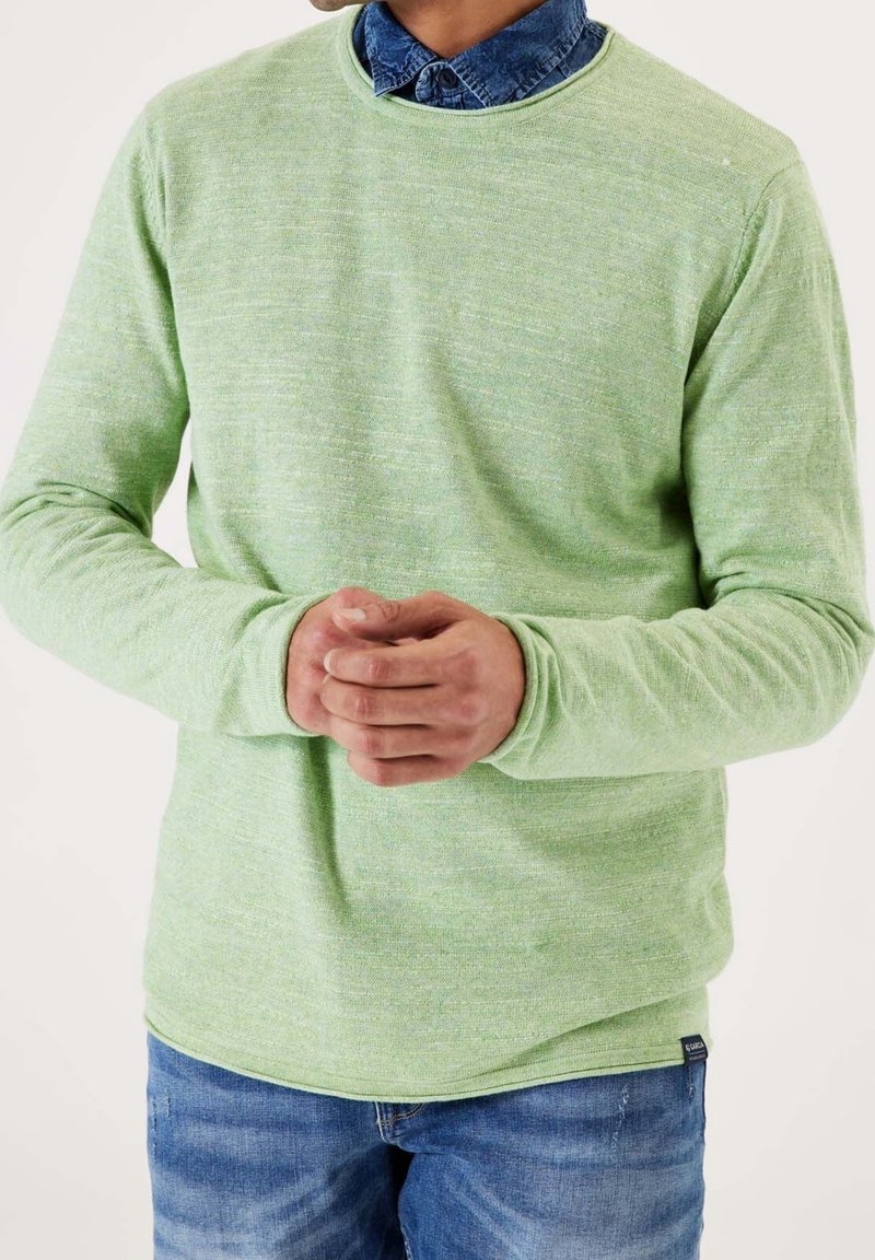 Homme portant un pull vert clair à manches longues par-dessus une chemise bleue à col et un jean bleu, mains jointes devant sur un fond uni.