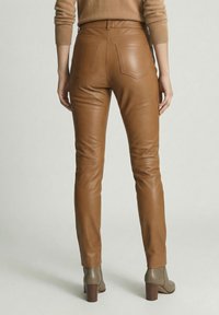 Vrouw draagt een bruine leren broek met hoge taille en achterzakken, gecombineerd met beige enkellaarsjes met hak, staand tegen een effen achtergrond.