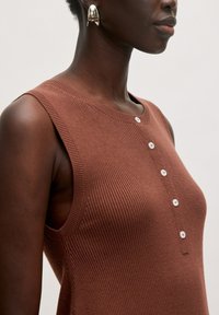 Bruine geribde tanktop met een ronde hals en witte knoopdetails aan de voorkant, gecombineerd met een glanzende zilveren oorbel.