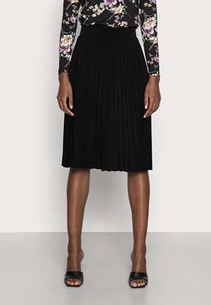 Femme portant une jupe plissée noire mi-genou, un haut à manches longues à fleurs, et des chaussures noires à bouts ouverts, debout devant un fond uni.