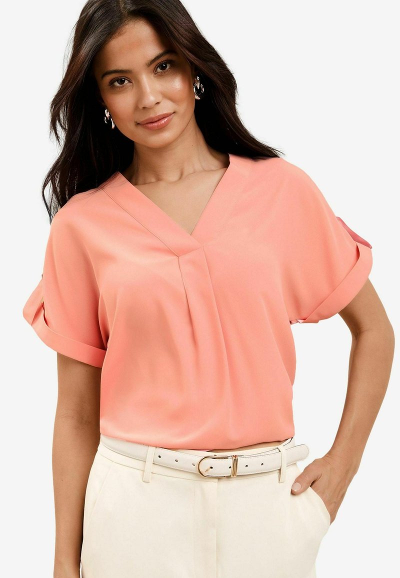 Blusa corta a maniche corte di colore rosa pallido con scollatura a V e maniche arrotolate, caratterizzata da delicate pieghe e abbinata a pantaloni a vita alta color crema e una cintura bianca.