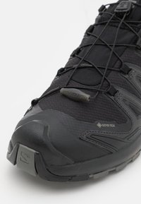 Black hiking shoe made of mesh and synthetic materials. Features a Gore-Tex label, rugged sole, and elastic lacing system for secure fit.