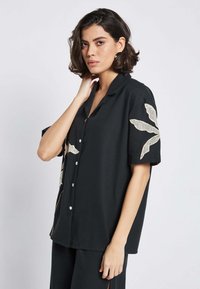 Maglietta nera a maniche corte con una vestibilità comoda. Presenta un'ampia ricamatura floreale beige sulle spalle e sul davanti, insieme a bottoni bianchi.