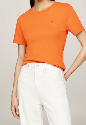 Kvinde iført en lyse orange kortærmet skjorte og højtaljede off-white bukser mod en ensfarvet baggrund.