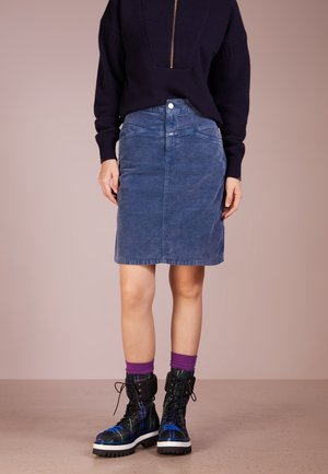 Femme portant un pull bleu marine, une jupe en jean bleue, des chaussettes violettes et des bottes noires à lacets à carreaux sur un fond beige uni.