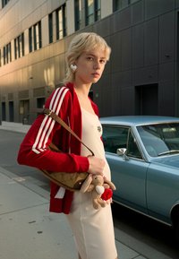 Veste rouge zippée avec des rayures blanches, robe crème, sac à main marron avec un motif matelassé, tenant un jouet en peluche lapin, debout à côté d'une voiture bleue.