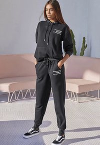 Schwarzes Hoodie- und Hosen-Set aus Baumwollmischung; mit Fronttasche, Kordelzug am Taillenbund und weißer Textdetail. Kombiniert mit schwarzen Sneakers.