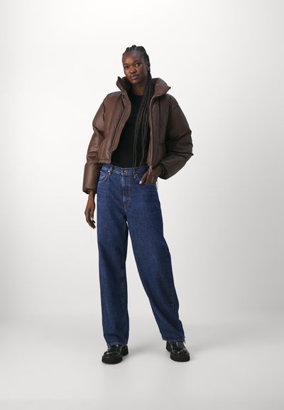 Doudoune marron avec col haut, pull en maille noir, jeans bleu à jambe large et chaussures noires chunky, se tenant contre un fond clair.