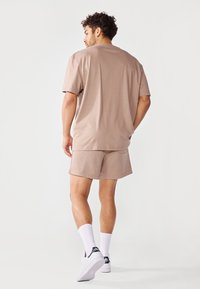 Beige katoenen t-shirt en short set, met een losse pasvorm. Zichtbare naden en stiksels. Gedragen met witte sokken en sneakers, met de rug naar de camera.