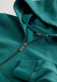 Zeleno-modra jopa z zadrgo, gladke teksture, nastavljivo kapuco in rebrastimi manšetami. Ima kontrastno zadrgo in etiketo na izrezu vratu.