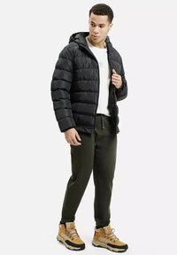 Czarna kurtka puchowa z kapturem, z poziomym pikowaniem. Noszona na białej koszuli, w zestawie z oliwkowymi spodniami i żółtymi butami.