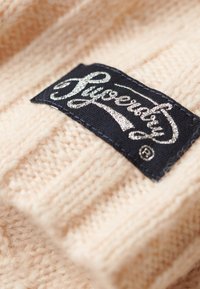 Tissu tricoté beige avec une texture côtelée, présentant une étiquette noire avec une écriture cursive blanche indiquant "Superdry". Vue en gros plan.