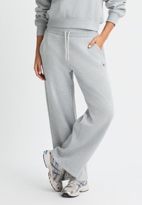 Pantalones de chándal gris claro con un ajuste relajado, cintura con cordón, bolsillos laterales y un pequeño detalle de logo en el muslo. Tela suave y textura lisa.