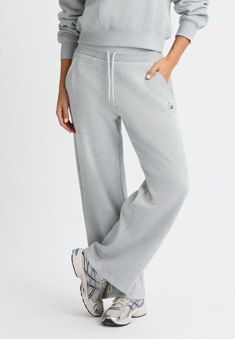 Pantalones de chándal gris claro con un ajuste relajado, cintura con cordón, bolsillos laterales y un pequeño detalle de logo en el muslo. Tela suave y textura lisa.