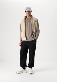 Beige fleece vest over a gray sweatshirt, paired with black sweatpants. Notable details include a zip pocket on the vest and light sneakers.