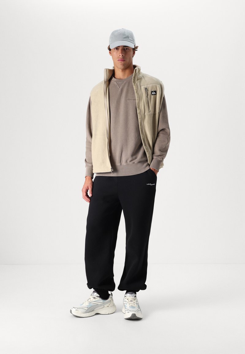 Beige fleece vest over a gray sweatshirt, paired with black sweatpants. Notable details include a zip pocket on the vest and light sneakers.